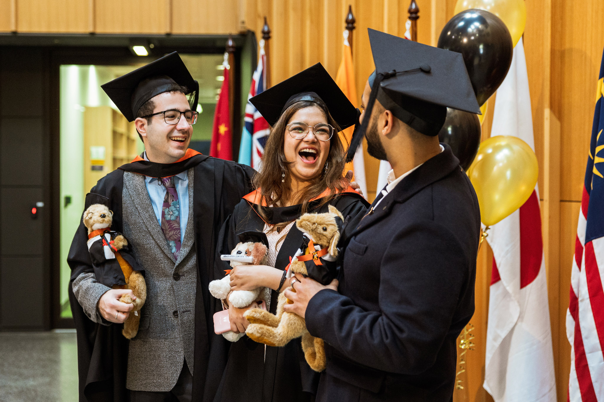 Students from the Coral Bell School of Asia Pacific Affairs celebrating at the post-graduation get together Three students wearing graduation gowns laughing together inside an esteemed-looking room, holding teddy bears