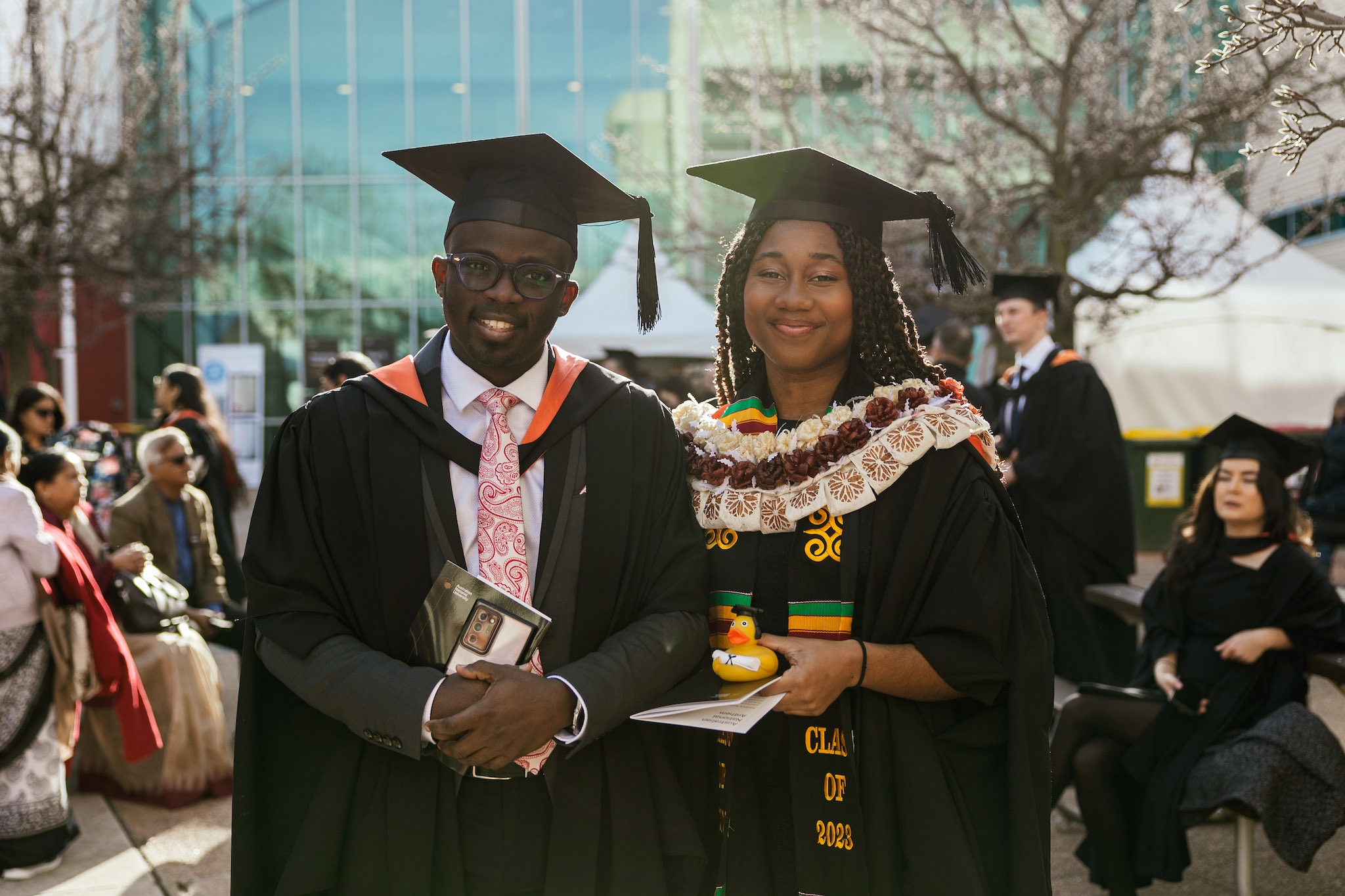 Graduate Adubi Plange (right) before the graduation ceremony outside Lewellyn Hall Two students smiling at the camera, wearing graduation gowns, standing outside Lewellyn Hall