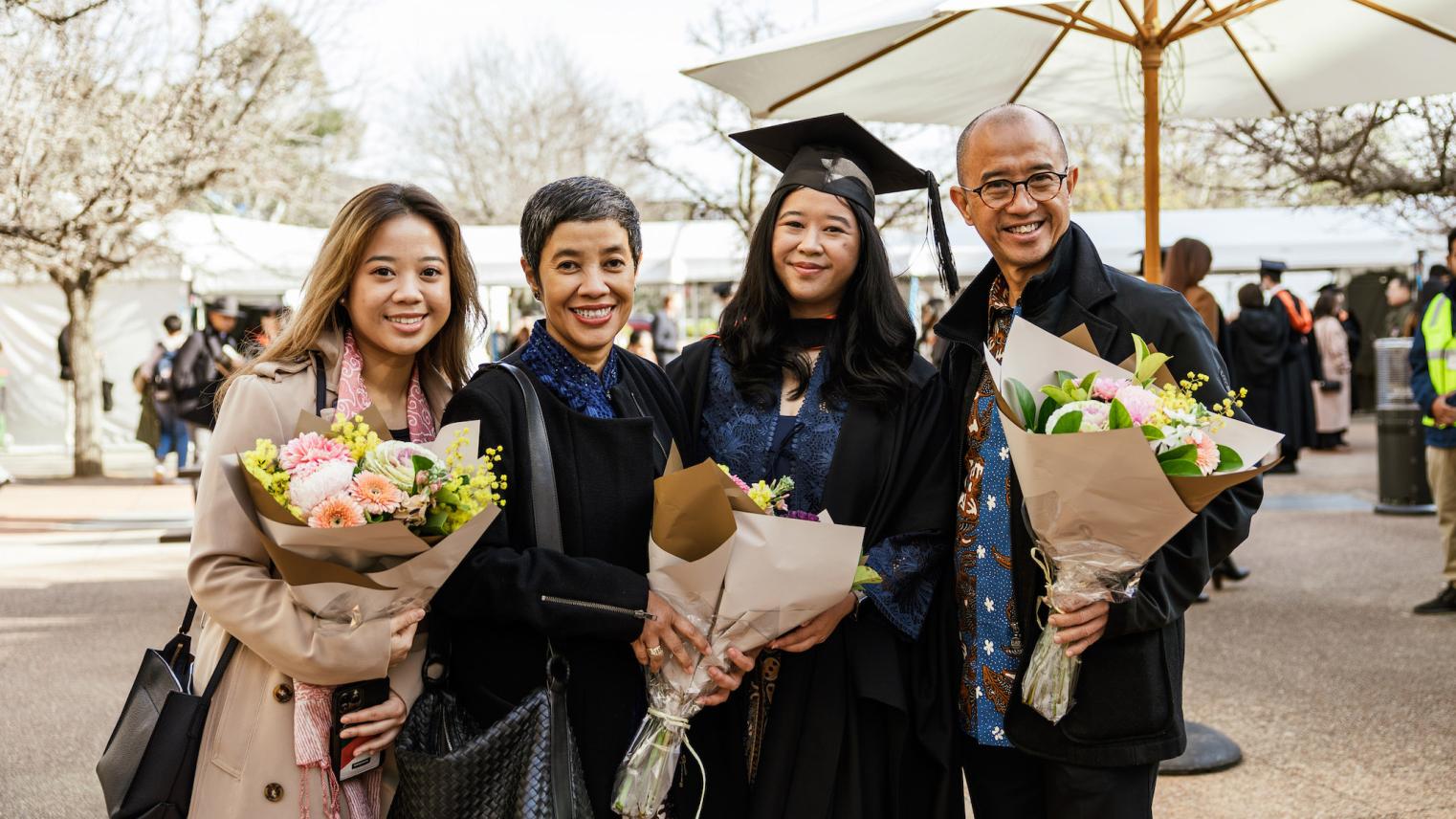 A family from the CAP graduations standing outdoors holding flowers and smiling A family from the CAP graduations standing outdoors holding flowers and smiling