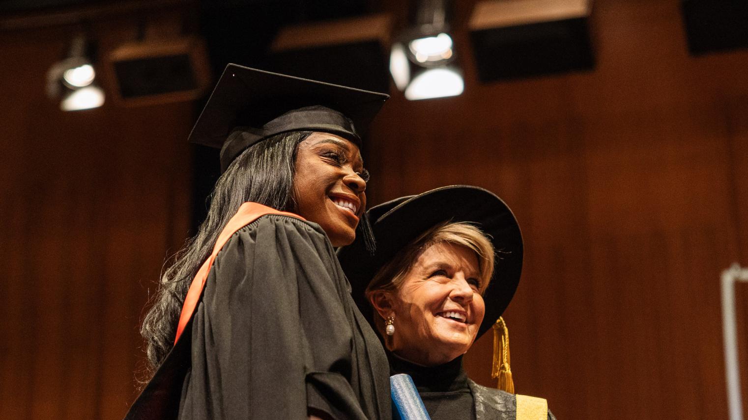 Precilla Anna-Kaii Lawrence standing on stage with Julie Bishop in graduation robes, holding her Testamur