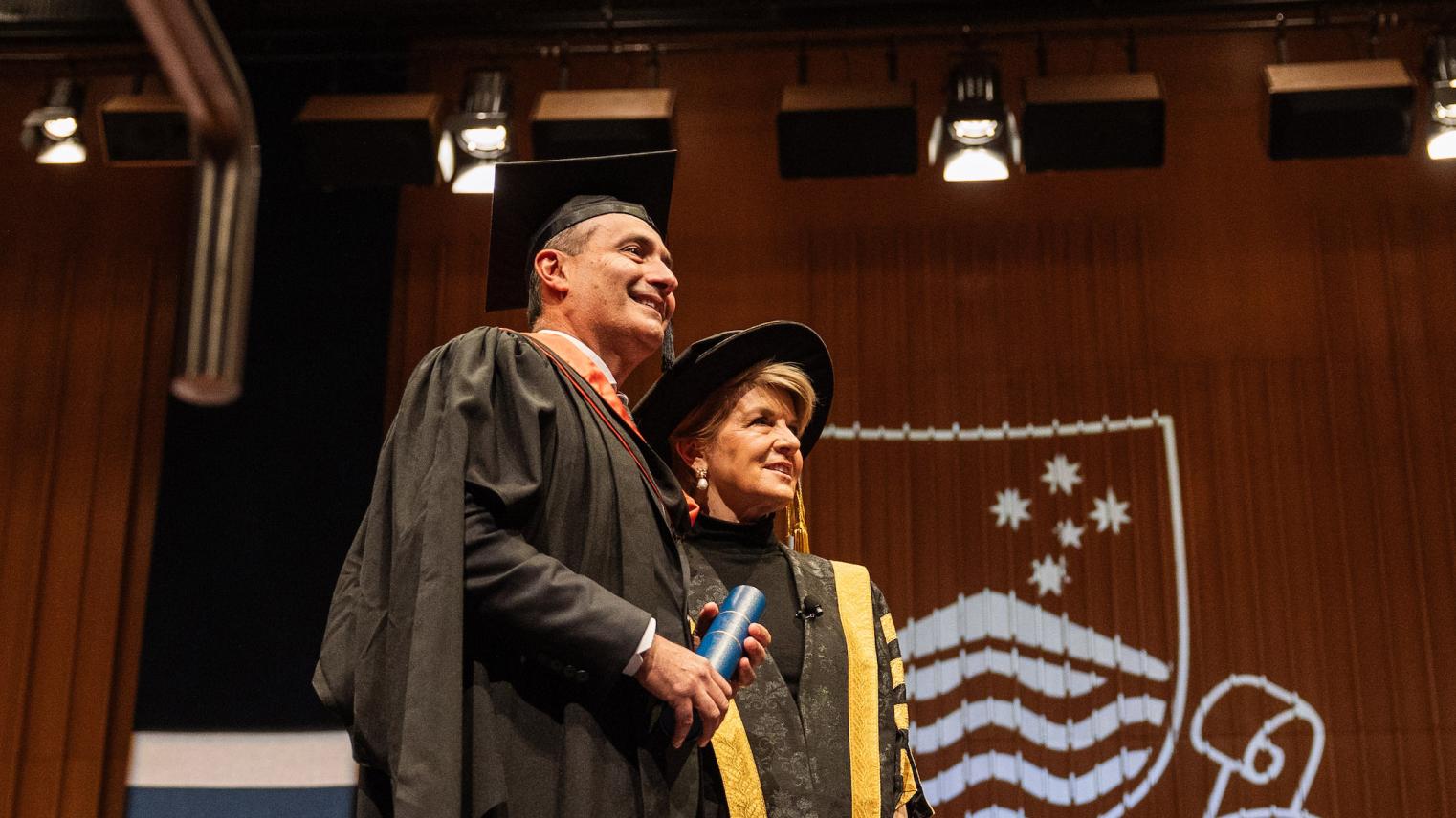 Steven Kourras standing on stage with Julie Bishop in graduation robes, holding his Testamur